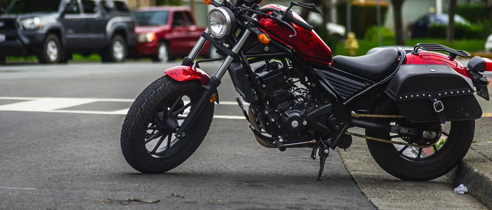 A red and black cruiser motorcycle with saddlebags parked at a street corner, with pickups parked behind